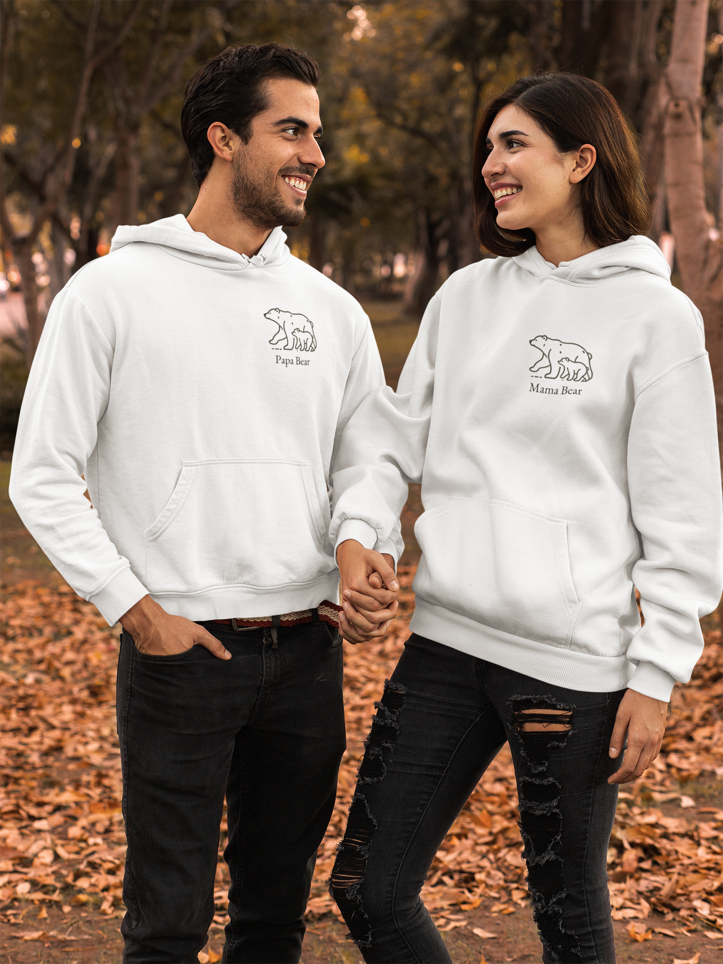 A couple holds hands outdoors with leaves all around them They are wearing matching white hoodies with their respective "Papa Bear" and "Mama Bear" designs on them and staring lovinginly at each other smiling